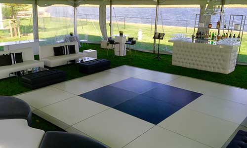 Photo of a portable outdoor dance floor in black and white. There is a tent canopy over the dance floor and there's  white bar and white couches in the background.  