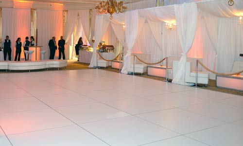 Photo of an event before the guests arrive: white portable dance floor with privacy booths in the back ground. There is a small stage for the band. 